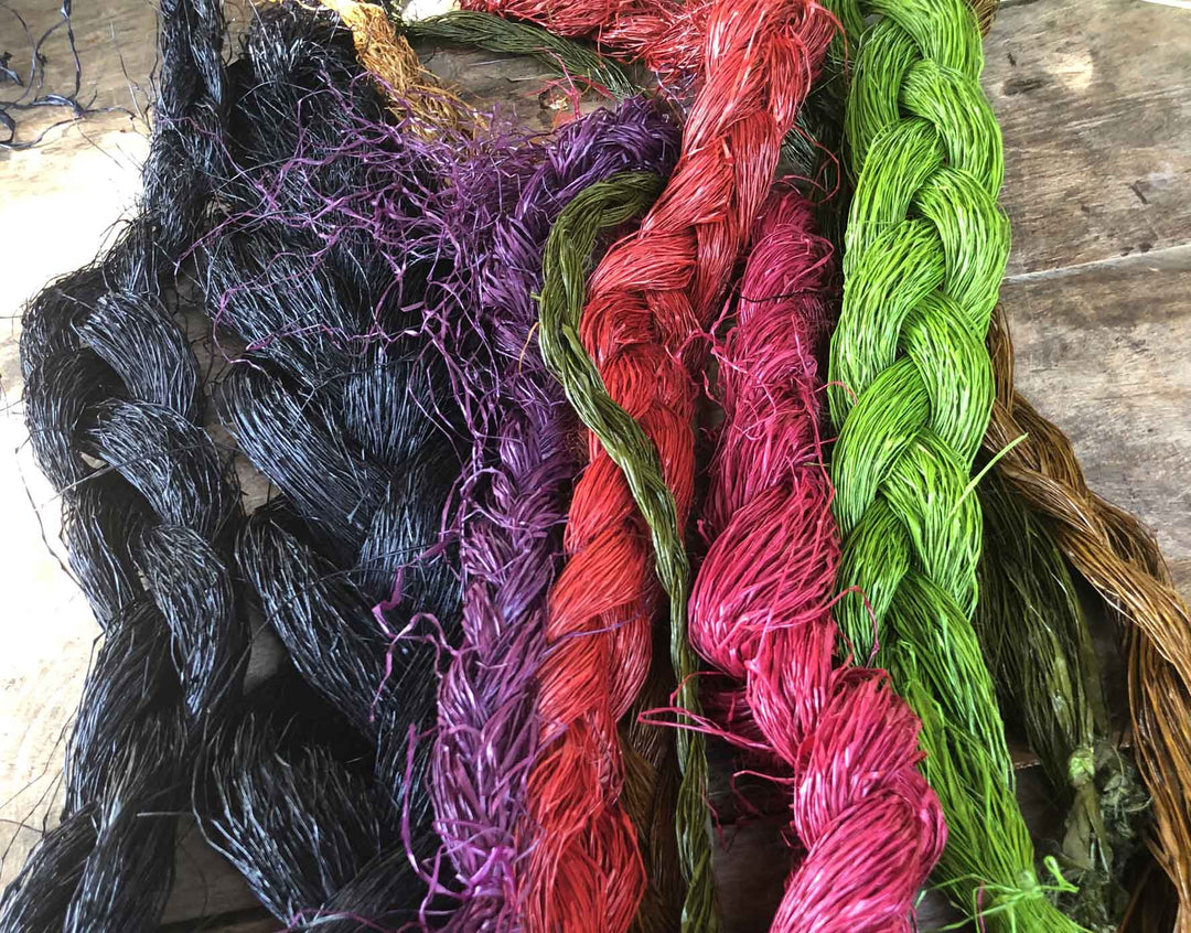 Braided hanks of hand-dyed chunga palm fiber in black, purple, red, pink, green and natural brown, laid side by side on a wooden table