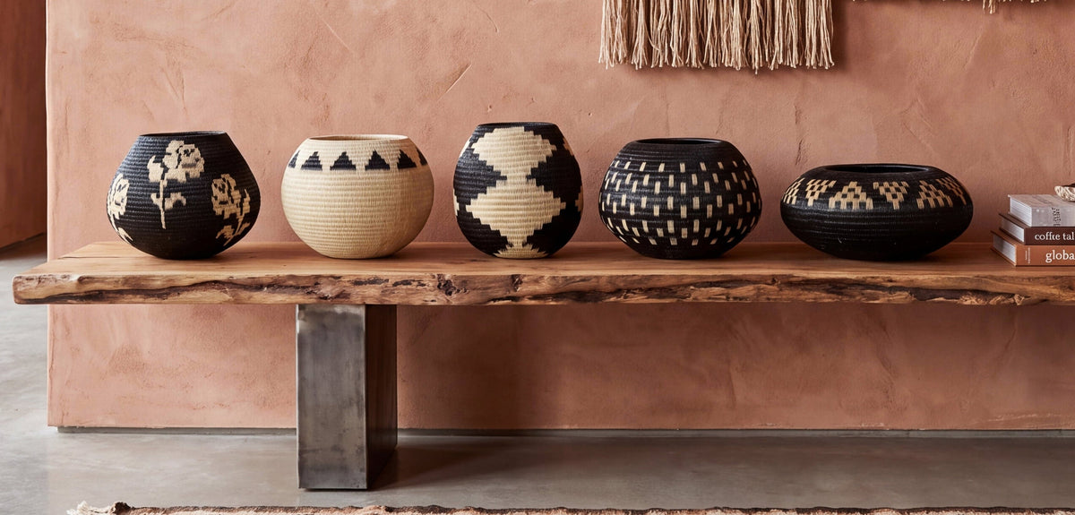 A styled interior scene showing a rustic wooden bench with several decorative woven baskets and a tall vase with dried grass, set against a terracotta wall with a woven wall hanging and rug.