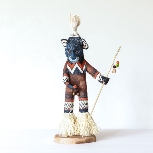 Ticuna spirit doll with a dark monkey face, chevron-painted body and raffia skirt, holding a beaded staff on a wood base
