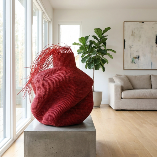 Sculptural red woven vessel with a fibrous fringe on a plinth in a bright living room