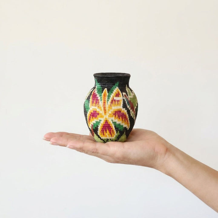 Mini Wounaan handwoven orchid basket held in hand showing miniature scale, gold and magenta bloom on black