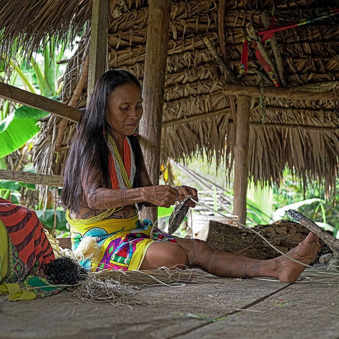 Woman weaving at a thatched hut with colorful fabric and natural surroundings