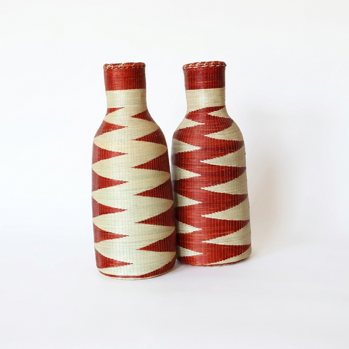 Pair of tall woven bottle vessels with red and cream zigzag bands on a white background