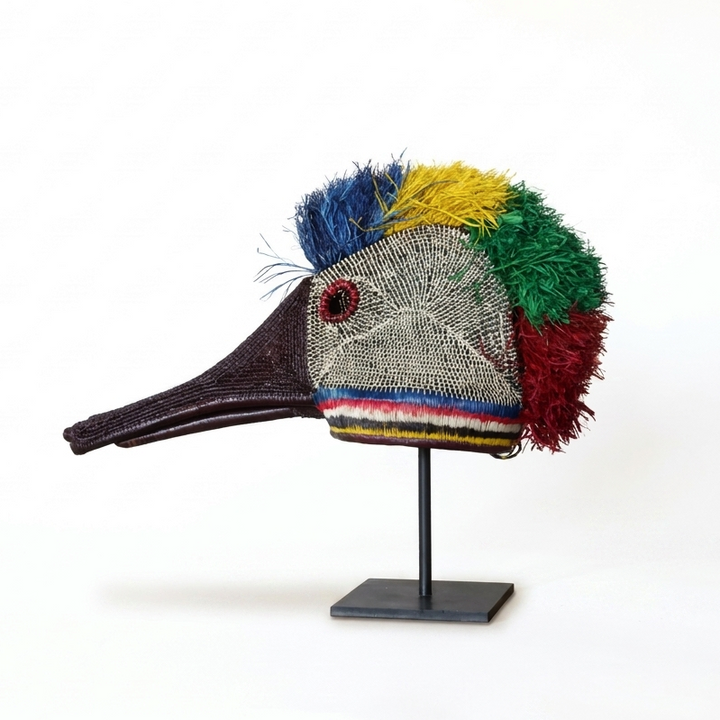 A decorative bird head sculpture or mask with a long dark beak, a gray mesh face, and colorful feather-like accents in blue, yellow, green, and red, displayed on a black stand against a plain white background.